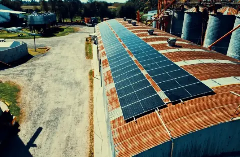 Paneles solares sobre el techo de un galpón largo, con silos al fondo, vista aérea